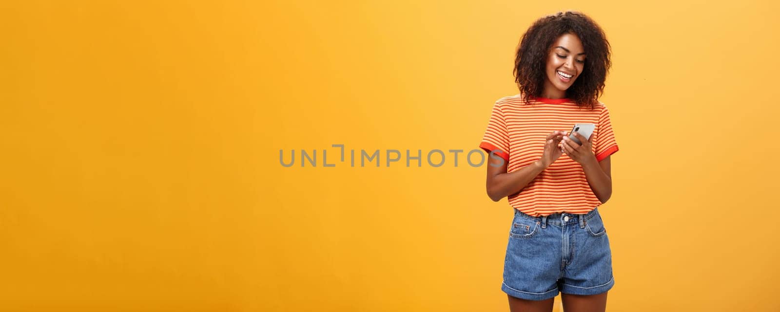 Stylish carefree girl texting friend come over standing pleased over orange wall in stylish denim shorts typing message or scrolling news in internet via smartphone gazing at device screen with smile by Benzoix