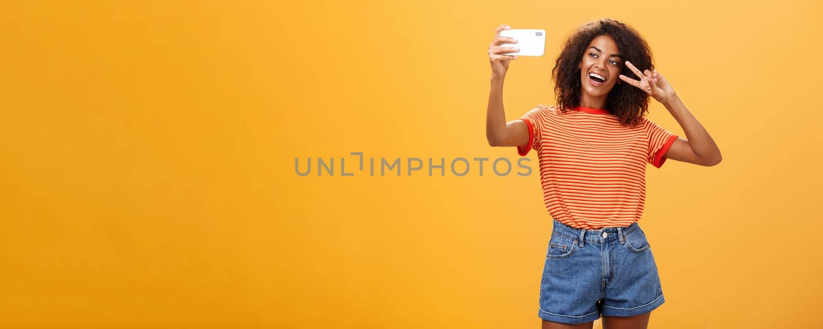 Girl making video vlog with brand new smartphone posting in internet trying become famous standing over orange background posing for selfie gazing at gadget screen showing peace or victory gesture. Copy space