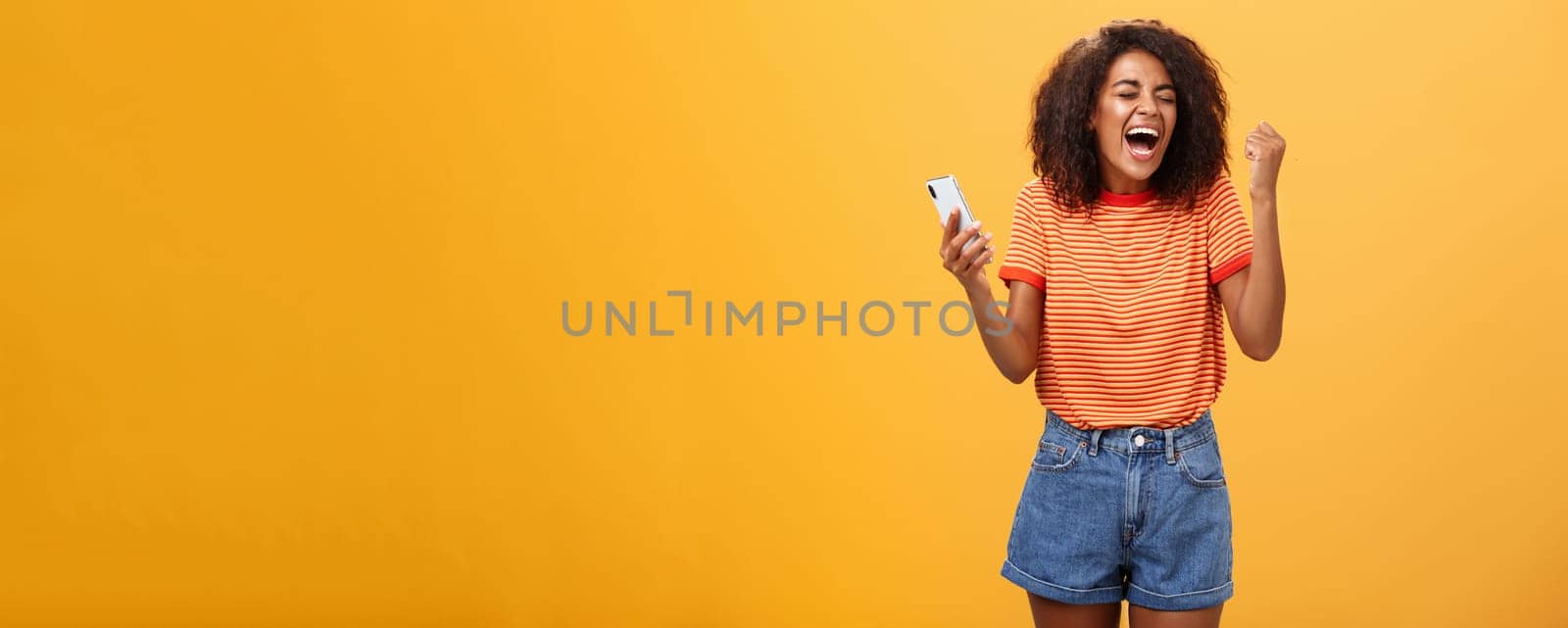 Portrait of ambitious happy young african american girl yelling from happiness and triumph clenching fist in joy and celebration feeling excited and relieved holding smartphone over orange wall by Benzoix