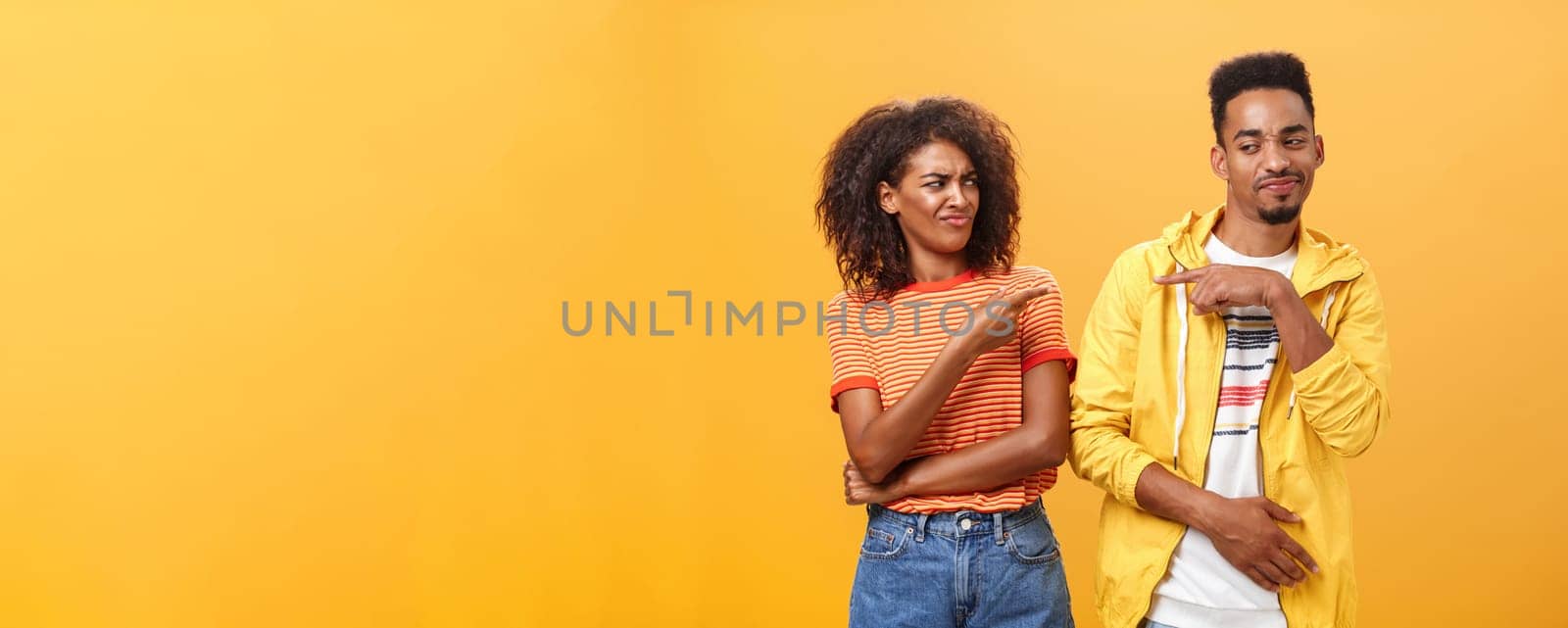 Two best friends fooling around having fun pointing at each other with cute joyful smile as if blaming each other playfully. Cute female looking at boyfriend standing together over orange wall.
