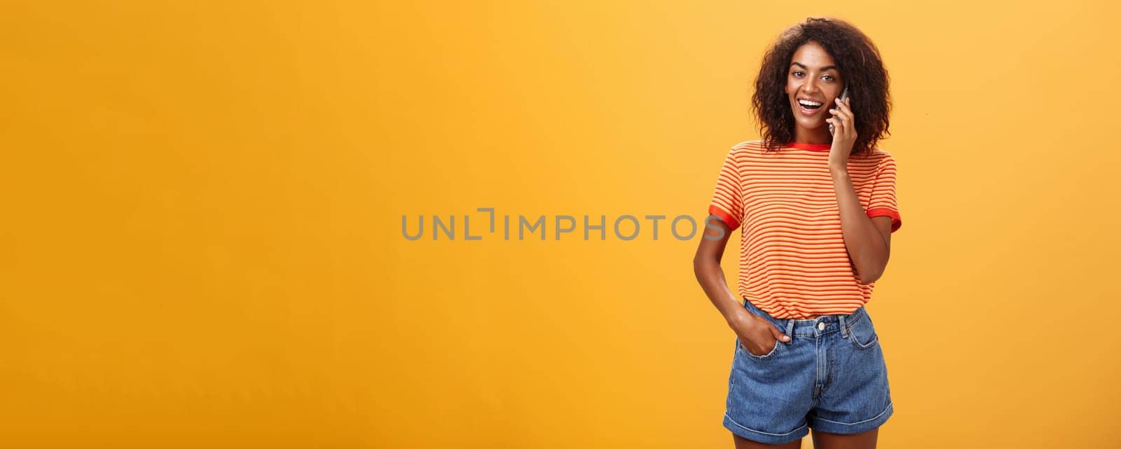 Indoor shot of stylish confident female coworker in trendy t-shirt and denim shorts holding smartphone near ear calling friend talking via cellphone casually about girls stuff over orange background. Lifestyle.