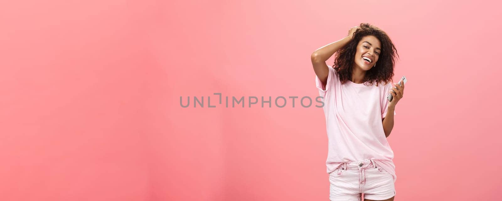 Portrait of carefree stylish modern dark-skinned young girl using smartphone hanging around joyfully touching hair and gazing with broad smile at camera holding cellphone posing against pink wall. Technology concept