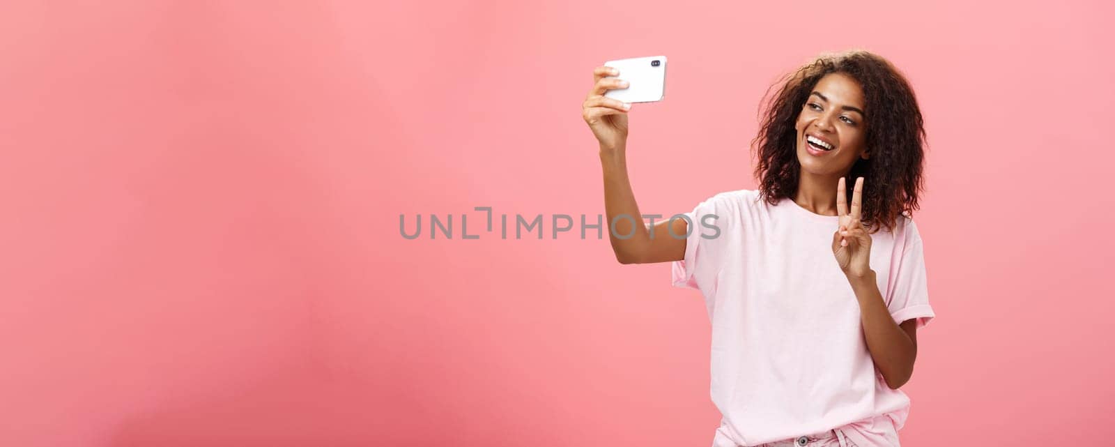 Stylish sociable good-looking dark-skinned female student with curly hairstyle pulling hand with smartphone near face taking selfie showing peace sign to device screen while smiling carefree. Lifestyle.