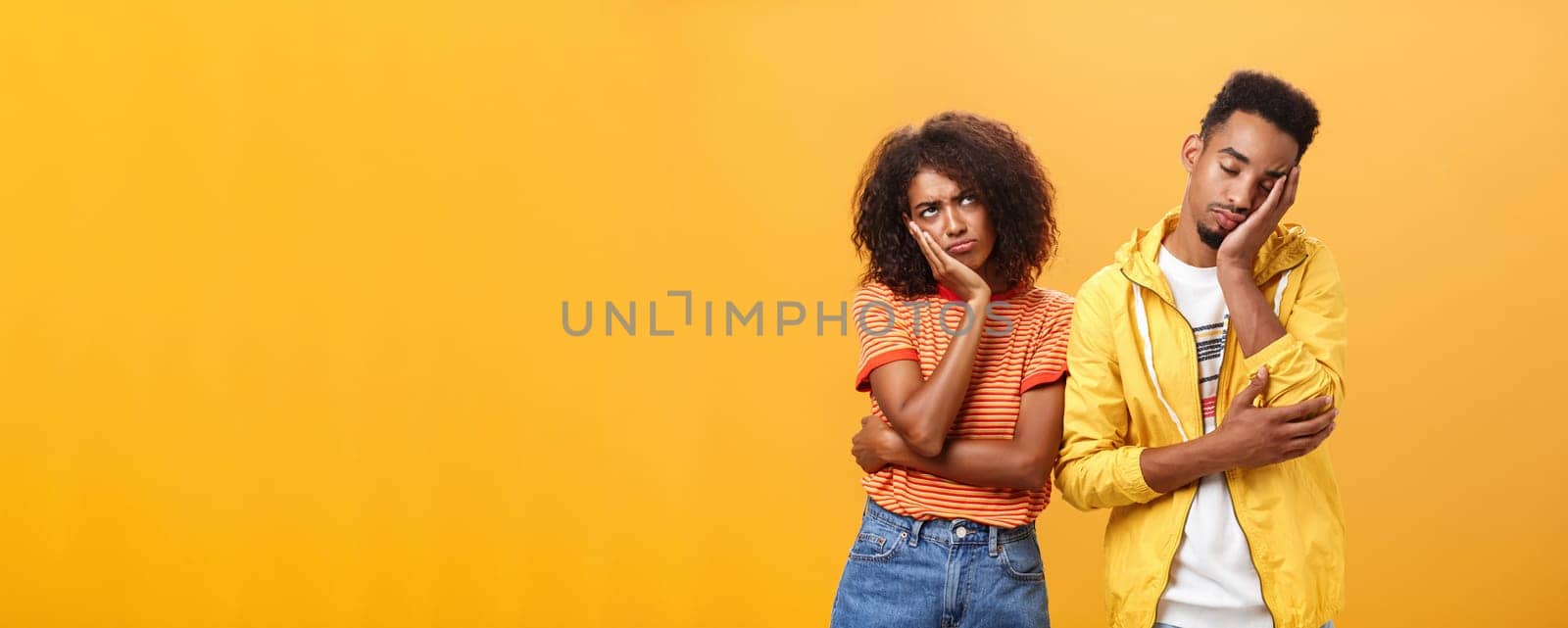 Girl feeling displeased and offended on boyfriend who fell asleep during date pursing lips frowning looking up while boyfriend leaning head on face with closed eyes and tired look over orange wall by Benzoix
