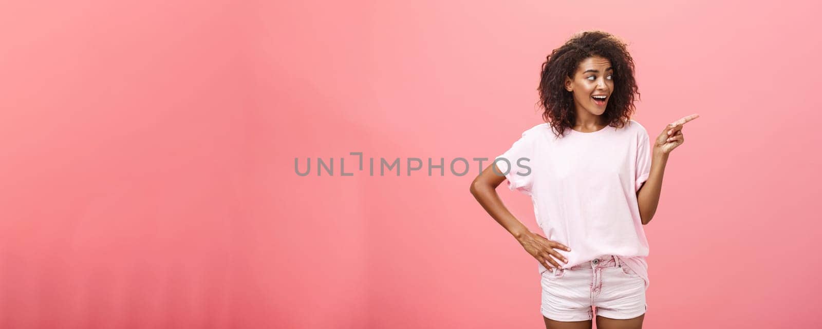 Hey wanna go there. Portrait of intrigued pleased good-looking stylish african woman on vacation wearing summer clothes pointing and gazing right with curious look over pink background. Advertisement concept