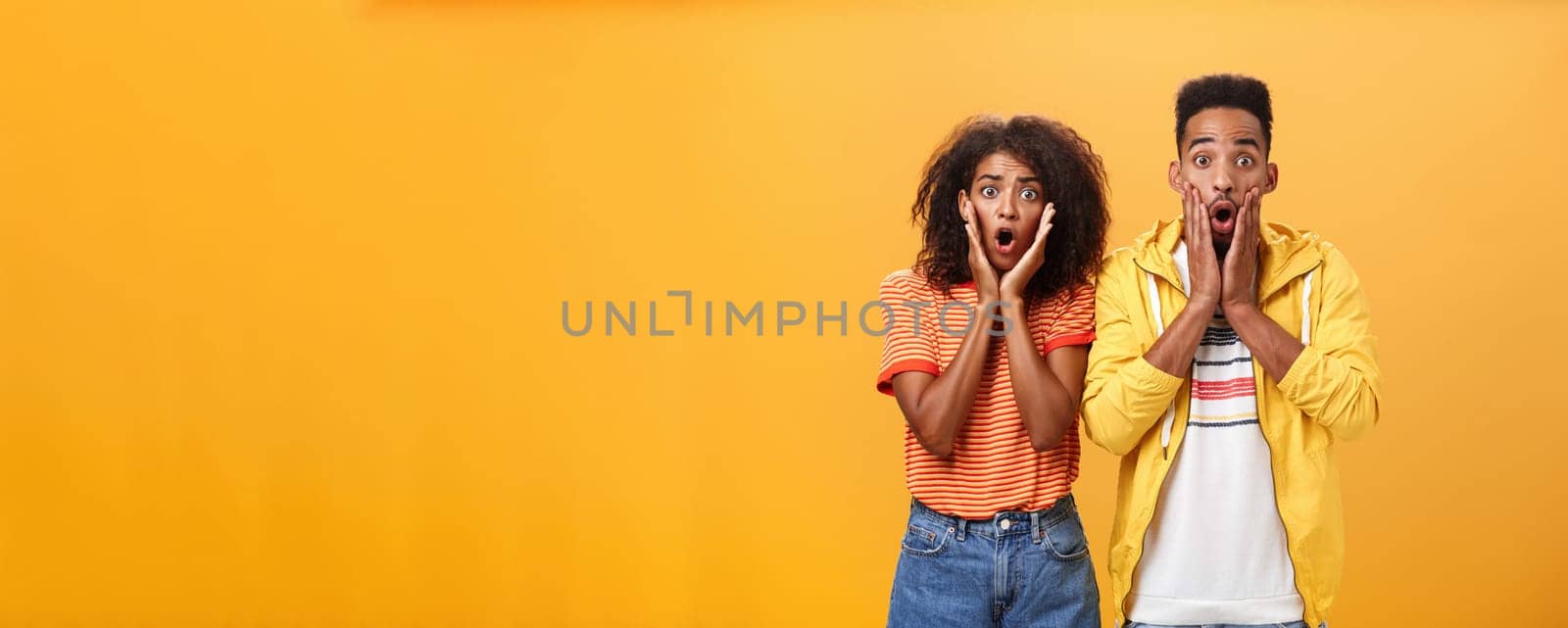 Portrait of shocked and stunned speechless girlfriend dropping jaw from amazement with boyfriend feeling amazed from shook news posing together surprised and astonished over orange wall. Lifestyle.