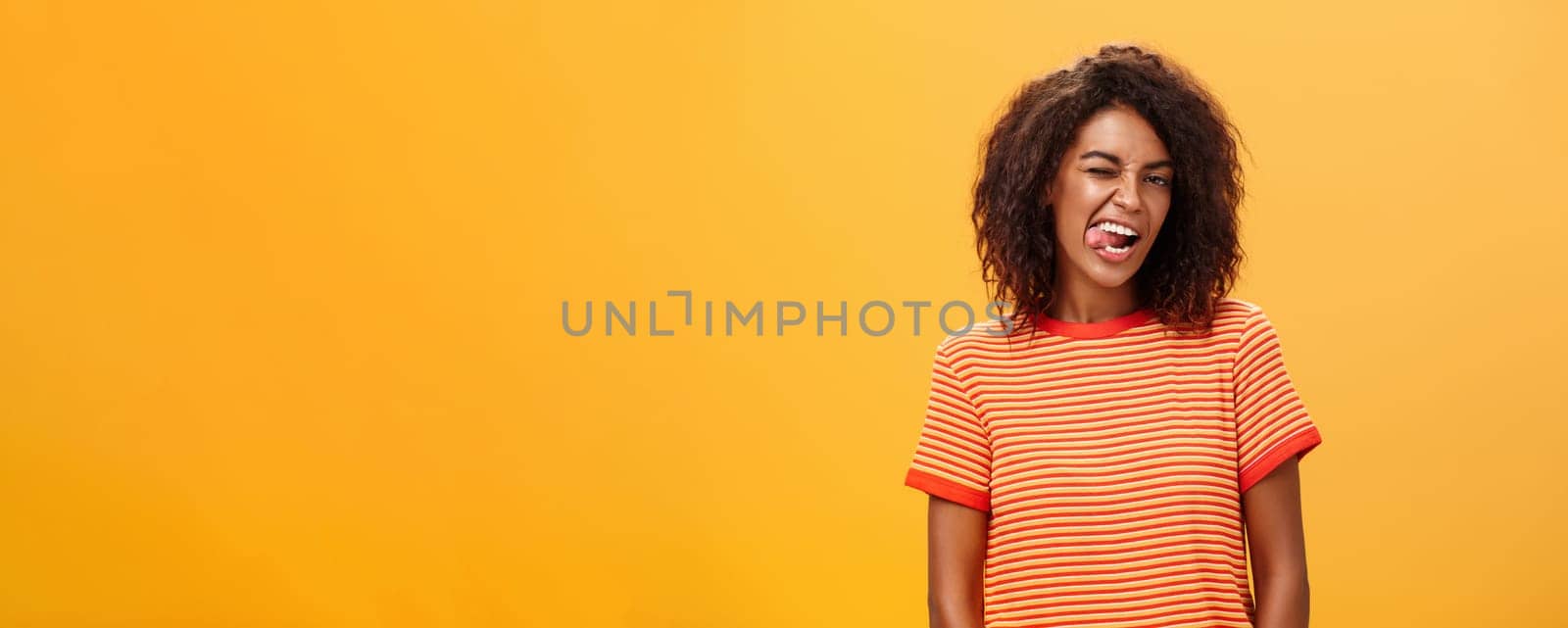Portrait of daring and emotive confident flirty woman with afro hairstyle winking joyfully showing tongue posing carefree and enthusiastic against orange background flirting with hot guy. Copy space
