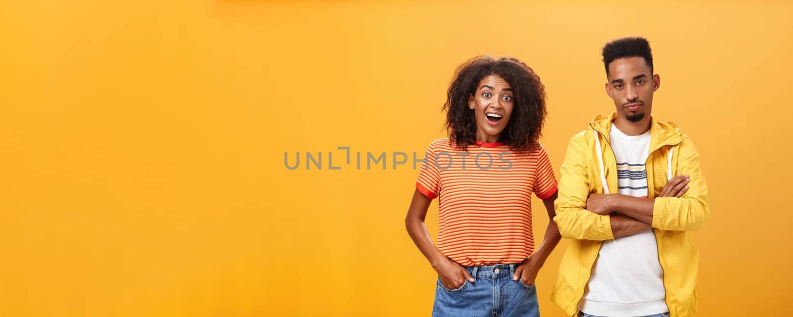 Two african american friends standing over orange background, girl thinks idea is awesome feeling excited and joyful while boyfriend being displeased and indifferent to unimpressive plan. Copy space