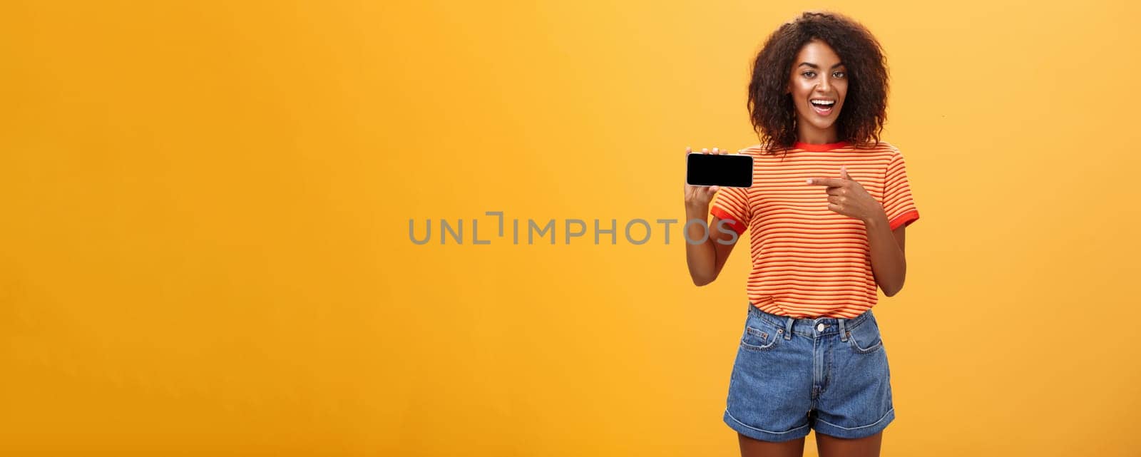 Girl showing picture in phone bragging about her cool vacation. Charming friendly and sociable african american woman with curly hair holding cellphone pointing at smartphone screen over orange wall. Technology and advertisement concept