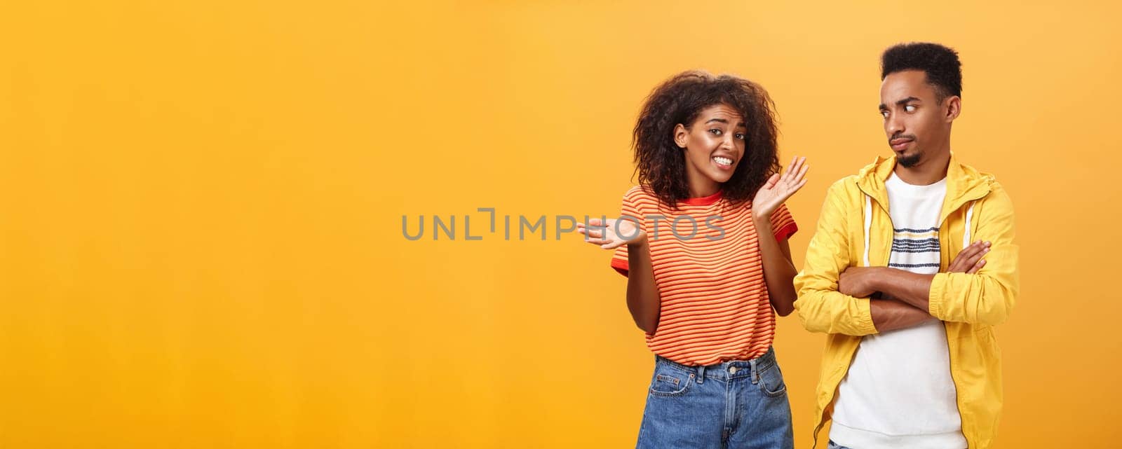 Guy thinks his friend weirdo making dumb thinks looking at cute girl with suspicious look crossing arms on chest raising eyebrow questioned while girlfriend saying sorry shrugging over orange wall by Benzoix