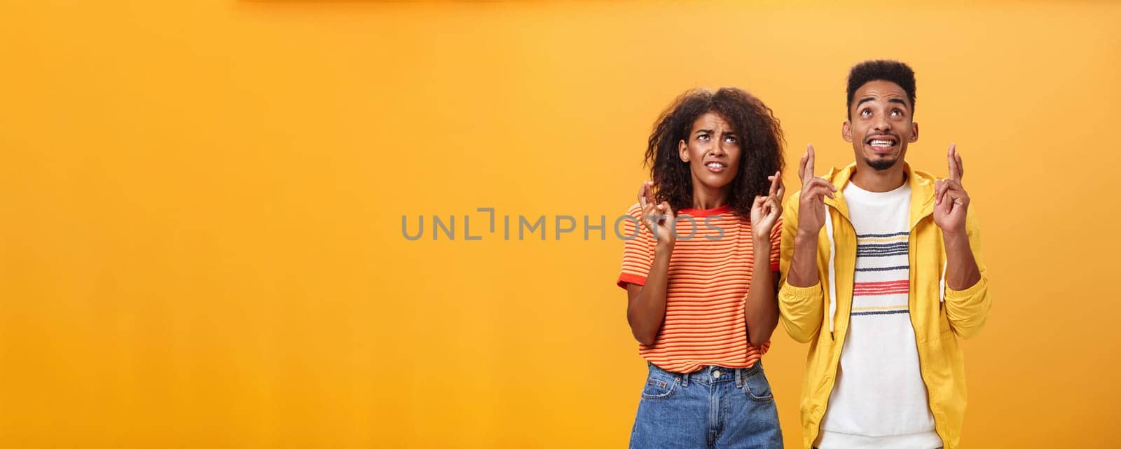 Couple of friends praying and looking up in sky intense crossing fingers for good luck clenching teeth faithfully hoping parents not saw them smoking standing together over orange background by Benzoix