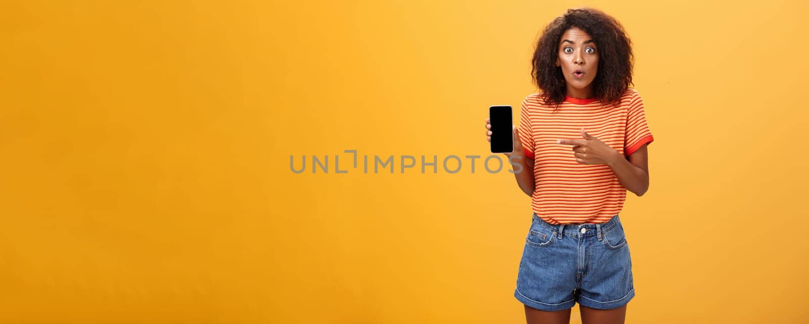 Portrait of impressed and curious female friend showing profile of ex to friend talking about shocking changes of appearance folding lips from interest holding smartphone pointing at cellphone screen. Lifestyle.