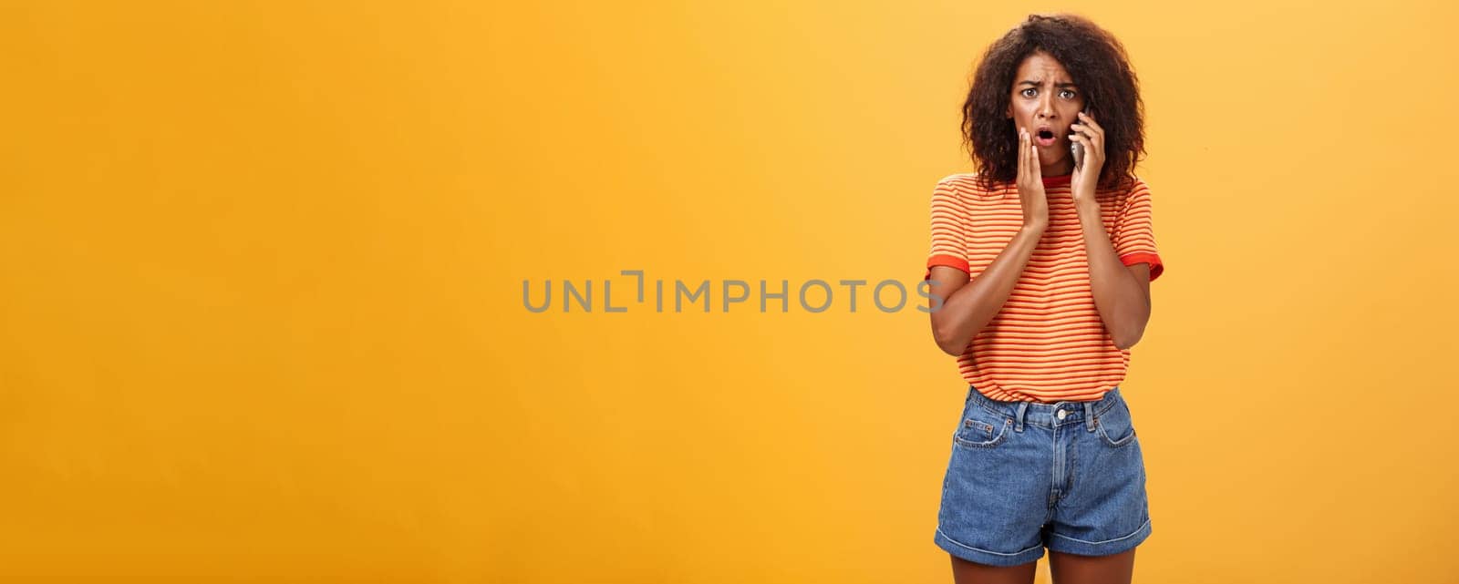 Woman receiving disturbing call feeling emapthy and sorry for poor friend getting in trouble holding cellphone near ear touching cheek concerned frowning standing sad, worried over orange background.