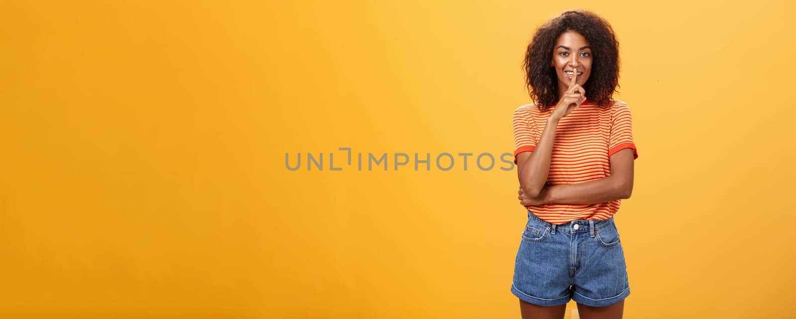 Keep secret inside I trust you. Feminine and sensual good-looking stylish dark-skinned woman with curly hair in trendy outfit shushing at camera with delighted mysterious look hiding something. Body language concept