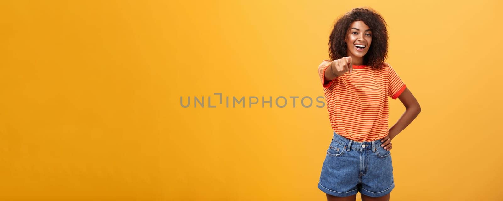 We hire you. Confident happy and awesome dark-skinned fashionable woman with curly hair holding hand on waist pointing at camera with index finger as if picking person or candidate, smiling. Body language concept