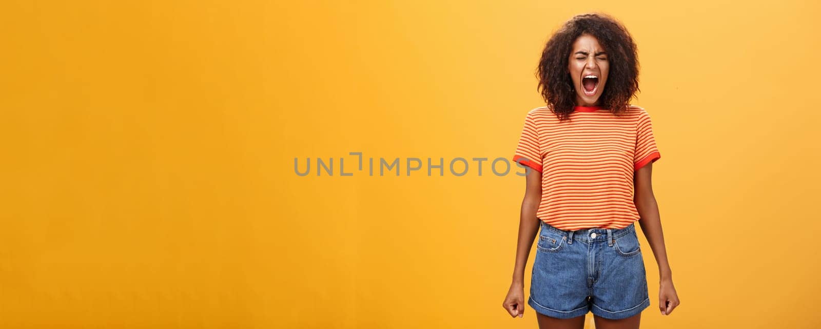 Girl feeling distressed and tensed losing temper and control of feelings yelling out loud clenching fists closing eyes standing intense and depressed over orange background screaming from rage by Benzoix