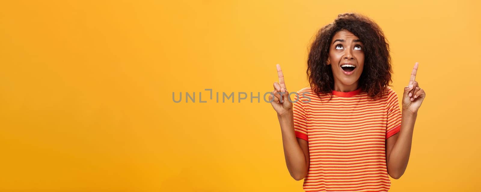 Optimistic happy african american girl saying thanks to god looking and pointing up pleased and carefree starting day in productive way being delighted with lucky chance over orange background. Body language and emotions concept