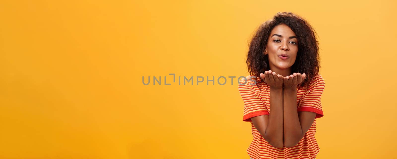 Sending passionate kiss to most loving person. Romantic attractive and stylish young african american girlfriend with curly hairstyle bending towards camera with slight smile, folded lips blowing mwah.