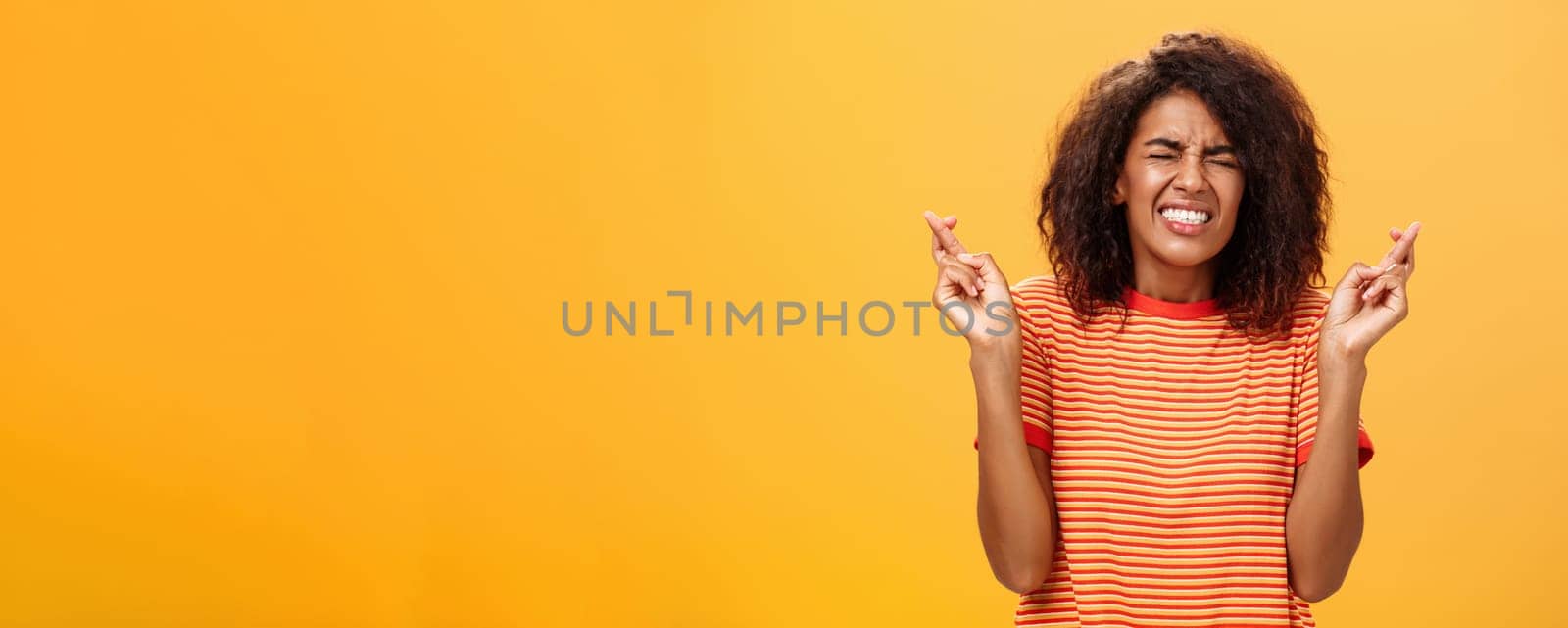 Waist-up shot of concerned worried woman eager take important job crossing fingers for good luck clenching teeth and closing eyes praying dreaming or making wish over orange background by Benzoix