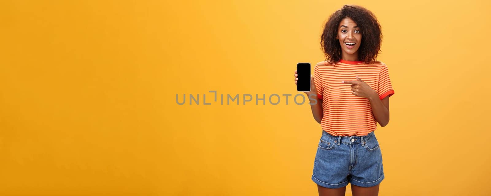 Cute african american stylish woman showing friend new app in smartphone pointing at cellphone screen with index finger smiling thrilled and happy delighted with cool device purchase over orange wall. Lifestyle.