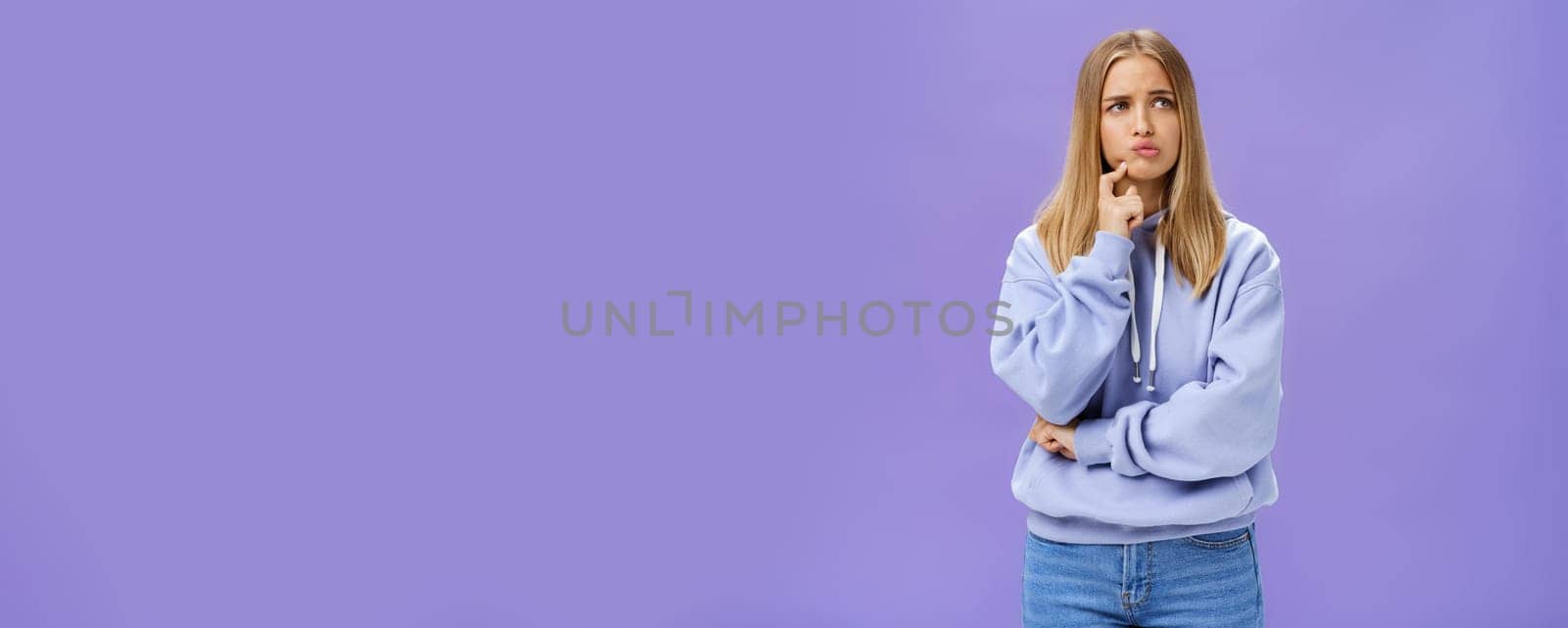 Silly concerned girlfriend trying think up how solve problem standing thoughtful with troubled expression touching jaw pouting, frowning staring right, thinking making up decision over purple wall. Body language and emotions concept