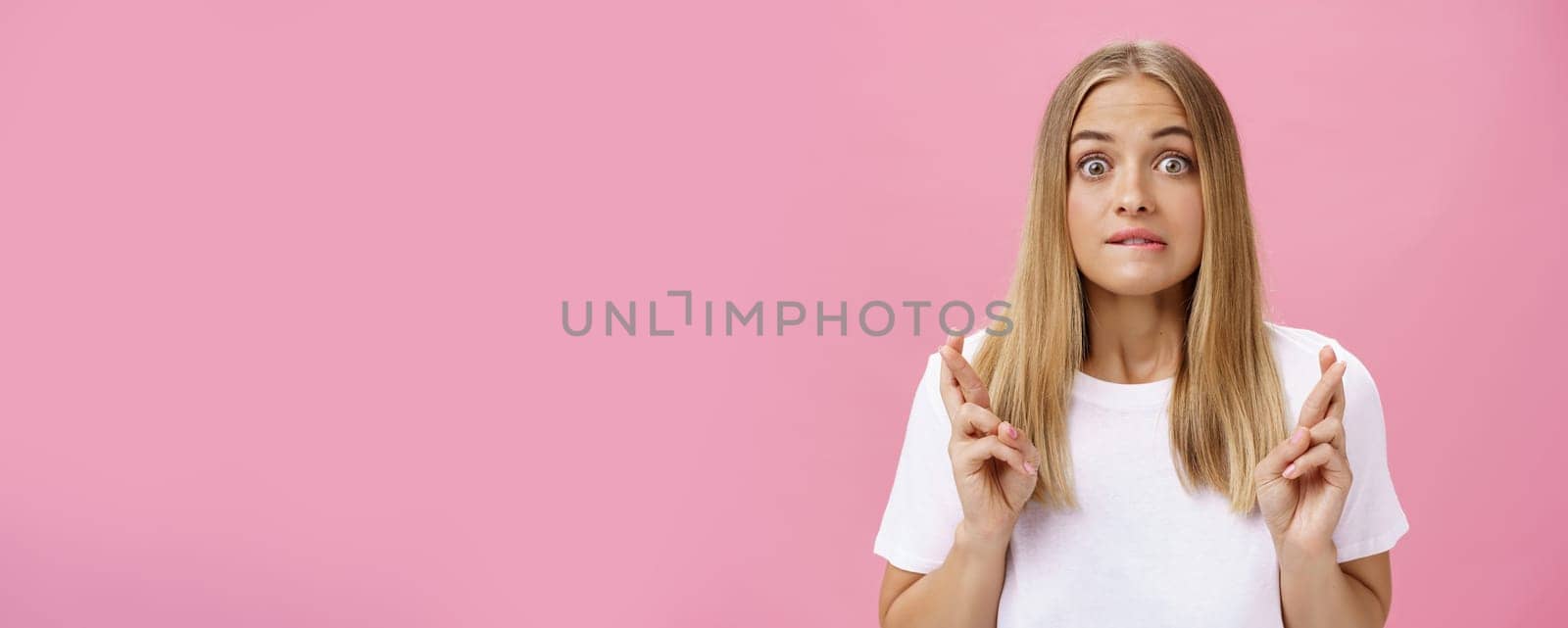 Excited and thrilled attractive emotive fair-haired female with pimples on forehead biting lower lips crossing fingers for good luck making wish praying desire become reality over pink wall. Emotions and body language concept