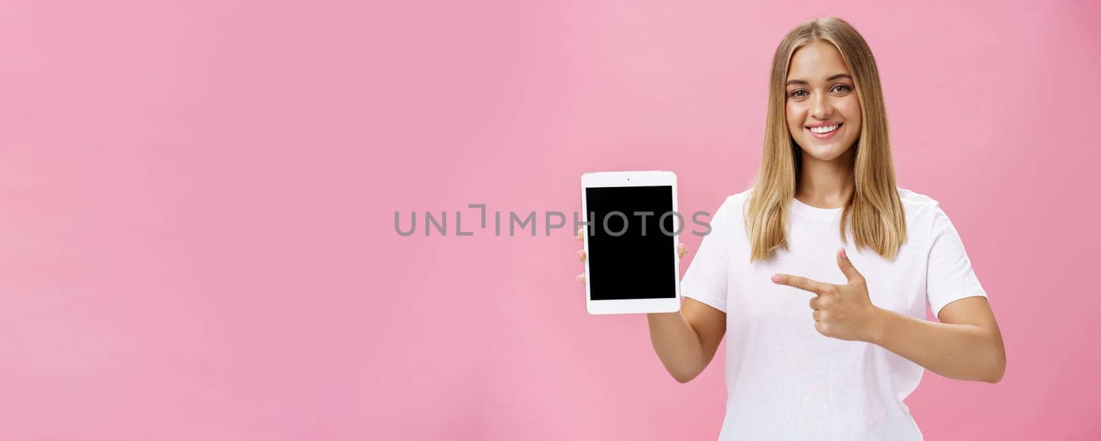 Girl recommends use only this digital tablet. Charismatic young female with fair hair tanned skin and no makeup in white t-shirt pointing at gadget screen and smiling friendly at camera over pink wall by Benzoix