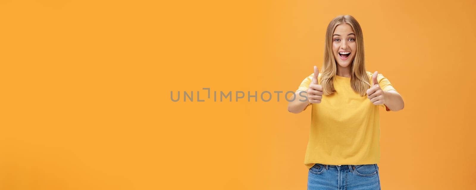 Woman supports with raised thumbs up and amused cheerful smile showing positive attitude expressing like on concept or idea giving approval posing happy and delighted against orange background. Lifestyle.