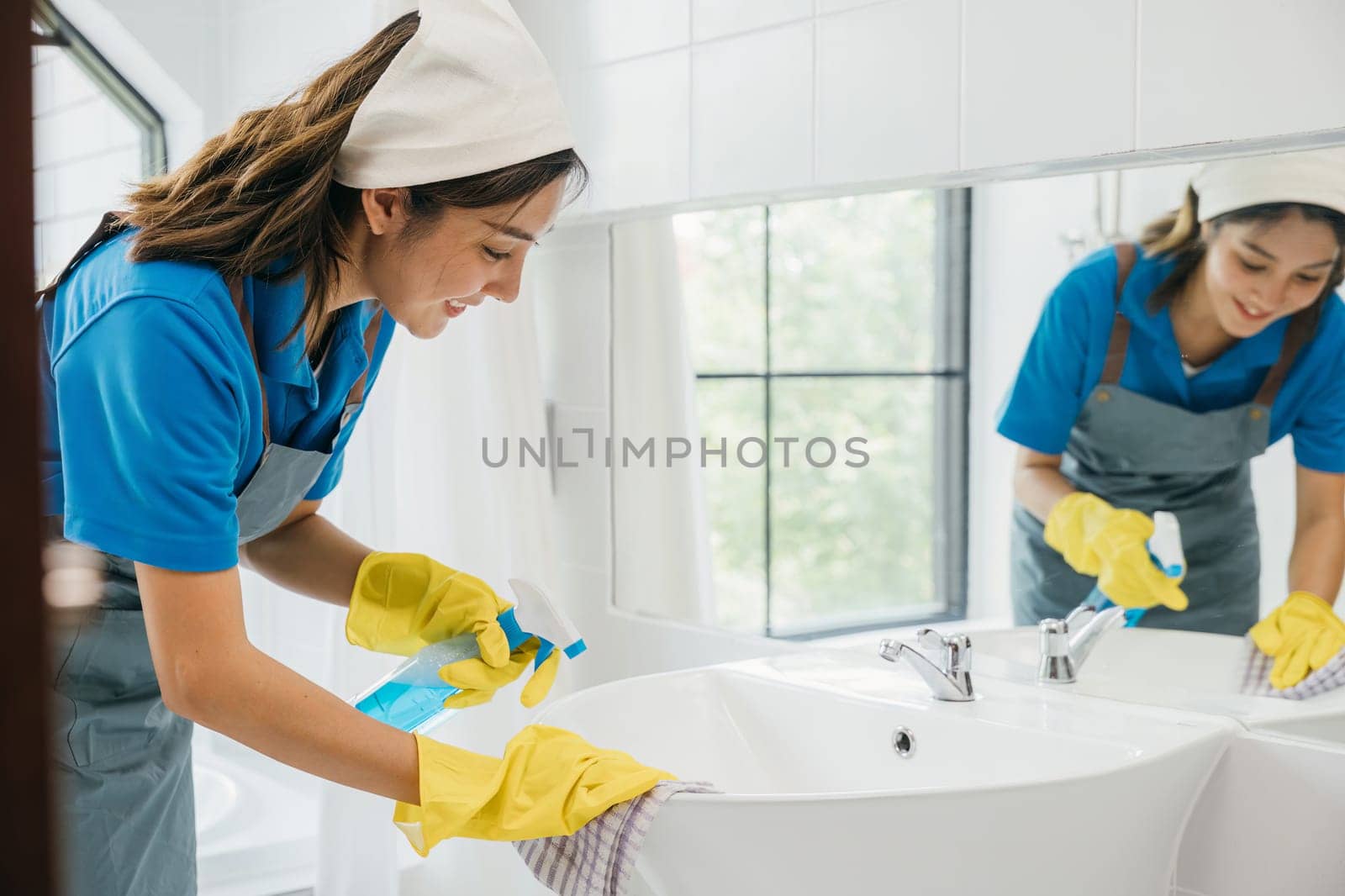 A maid wearing gloves diligently cleans bathroom sink and faucet eliminating limescale with spray detergent. Her focus on housework ensures purity hygiene and shining fixtures. spray cleaner by Sorapop