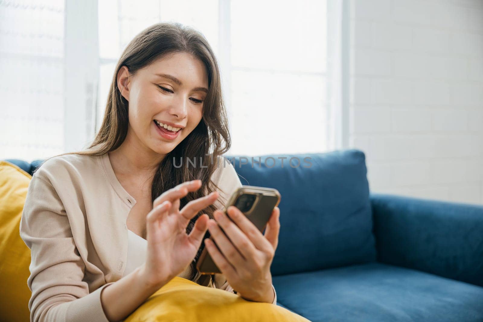 In a relaxed home setting a woman on a sofa happily talks on her phone typing messages. Engaged in successful communication and technology she embodies relaxation. The woman is relaxing.