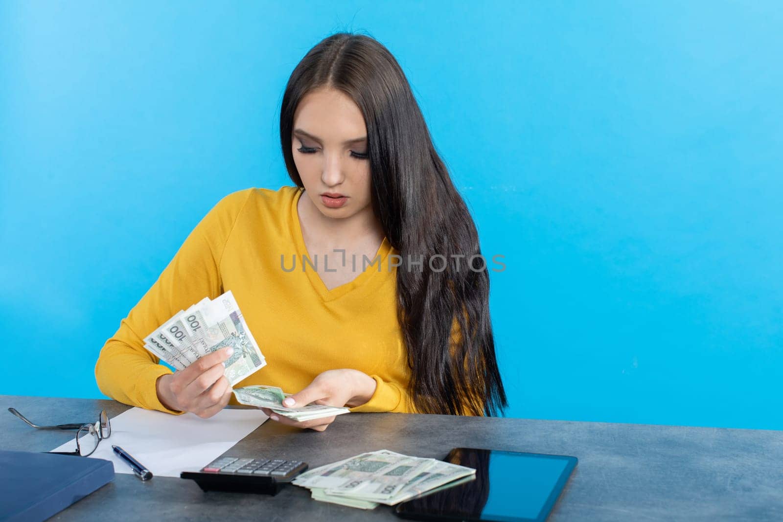 While counting money, a young woman uses a calculator and a tablet to check the stock exchange.