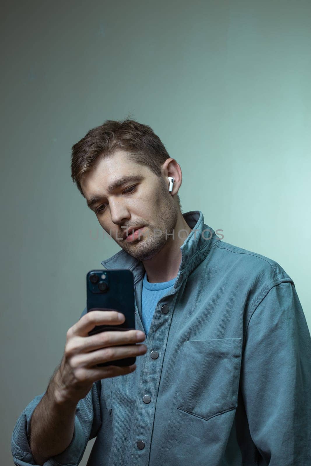 A thoughtful man in a blue shirt focused on his cellphone against a plain backdrop, immersed in technology