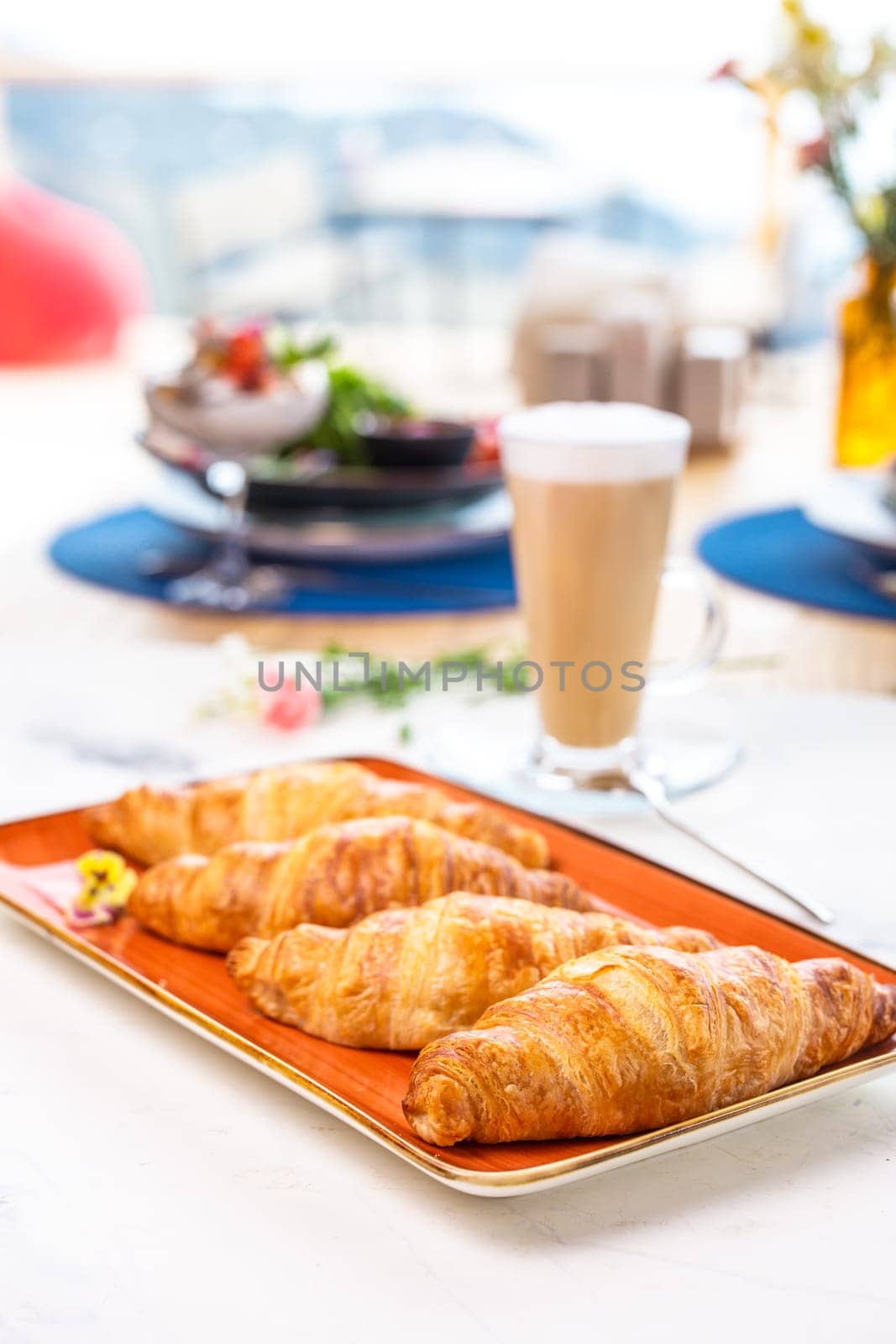 A delightful breakfast with croissants and coffee on a marble table. Croissants are golden brown on an orange plate, coffee with froth.