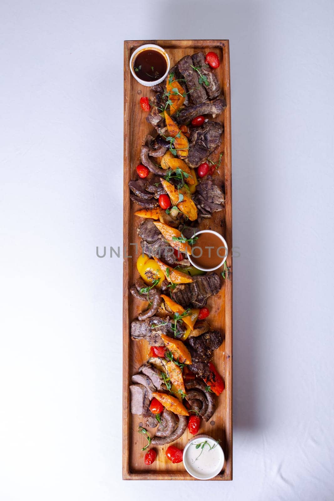 A long wooden platter filled with an assortment of meats and vegetables, including steak, sausage, chicken, potatoes, carrots, and peppers by Pukhovskiy