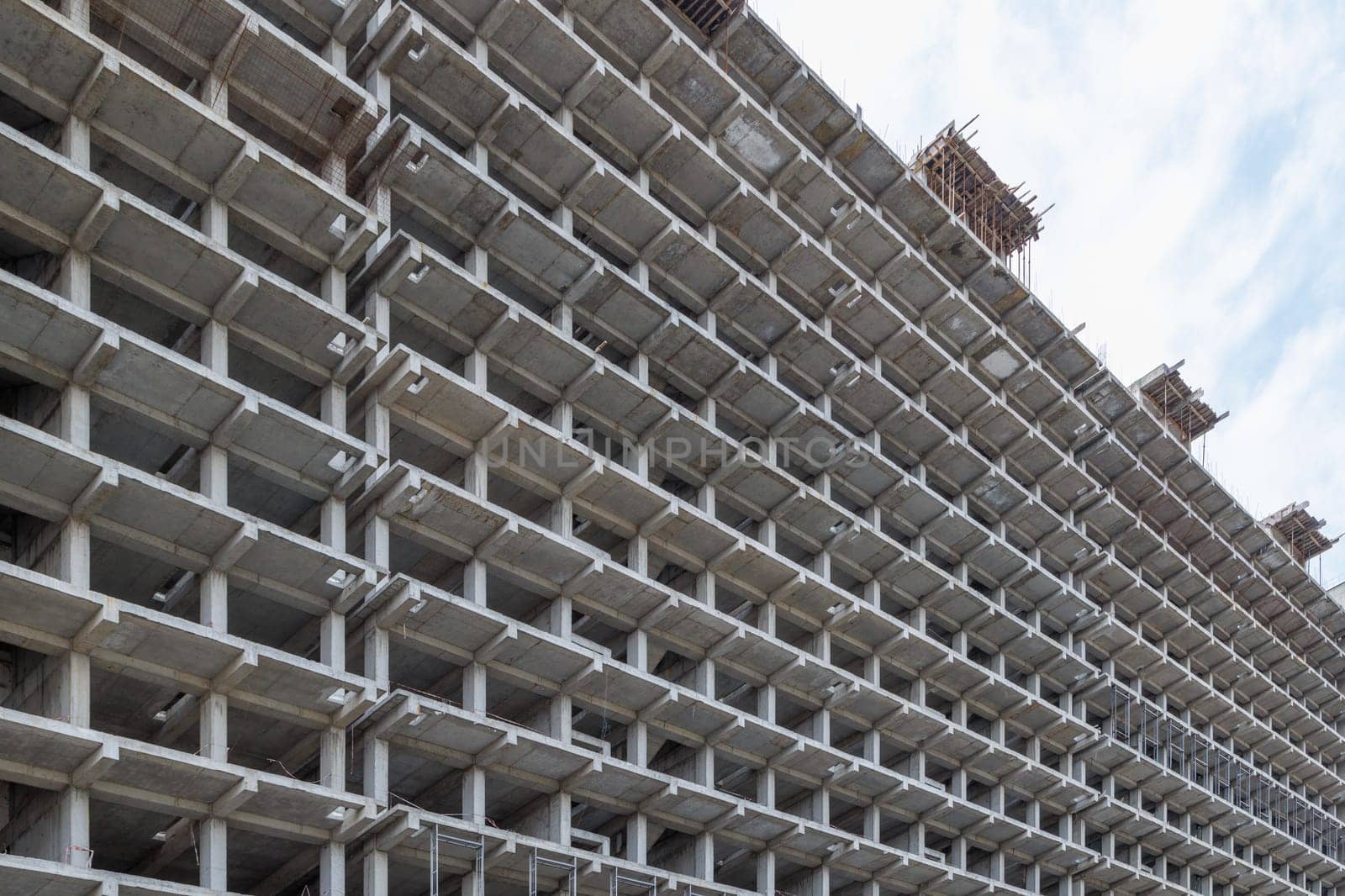 A large multi-storey apartment building is under construction, with a grey facade and a pattern of holes. The engineering fixtures are visible against the cloudy sky