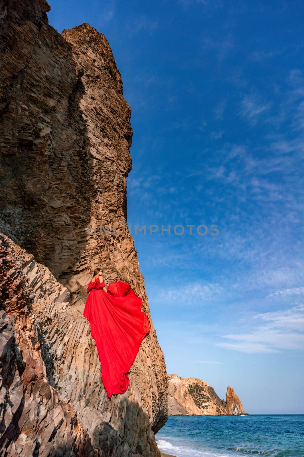 woman sea red dress. Woman with long hair on a sunny seashore in a red flowing dress, back view, silk fabric waving in the wind. Against the backdrop of the blue sky and mountains on the seashore. by Matiunina