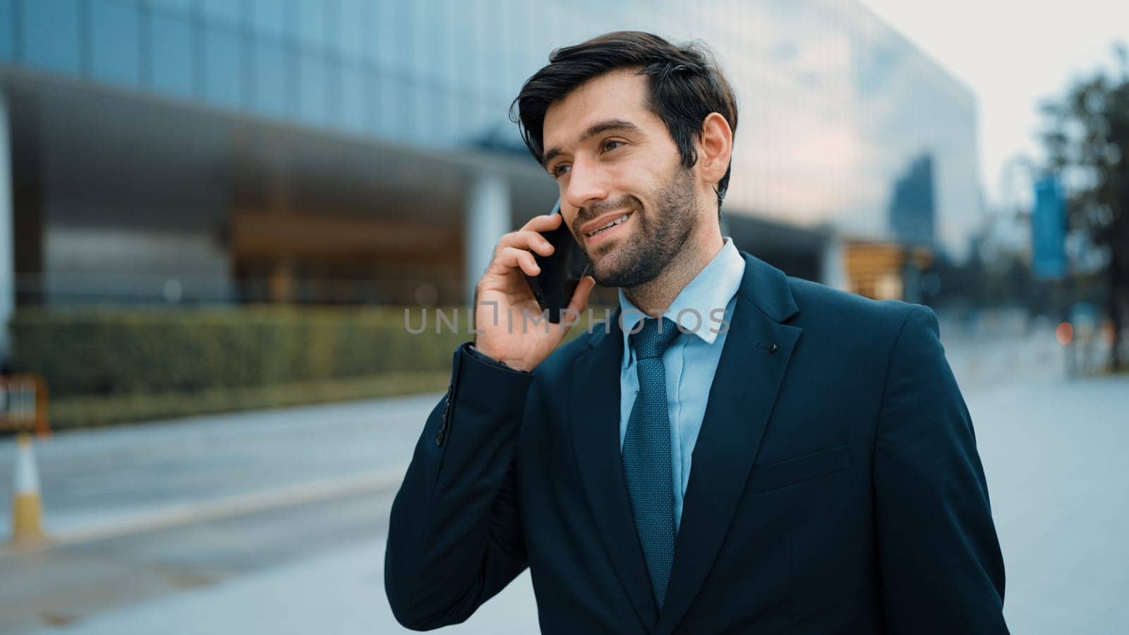 Skilled caucasian project manager calling phone while walking at city with blurred background. Skilled businessman using smart phone talking to project manager with blurred background. Exultant.