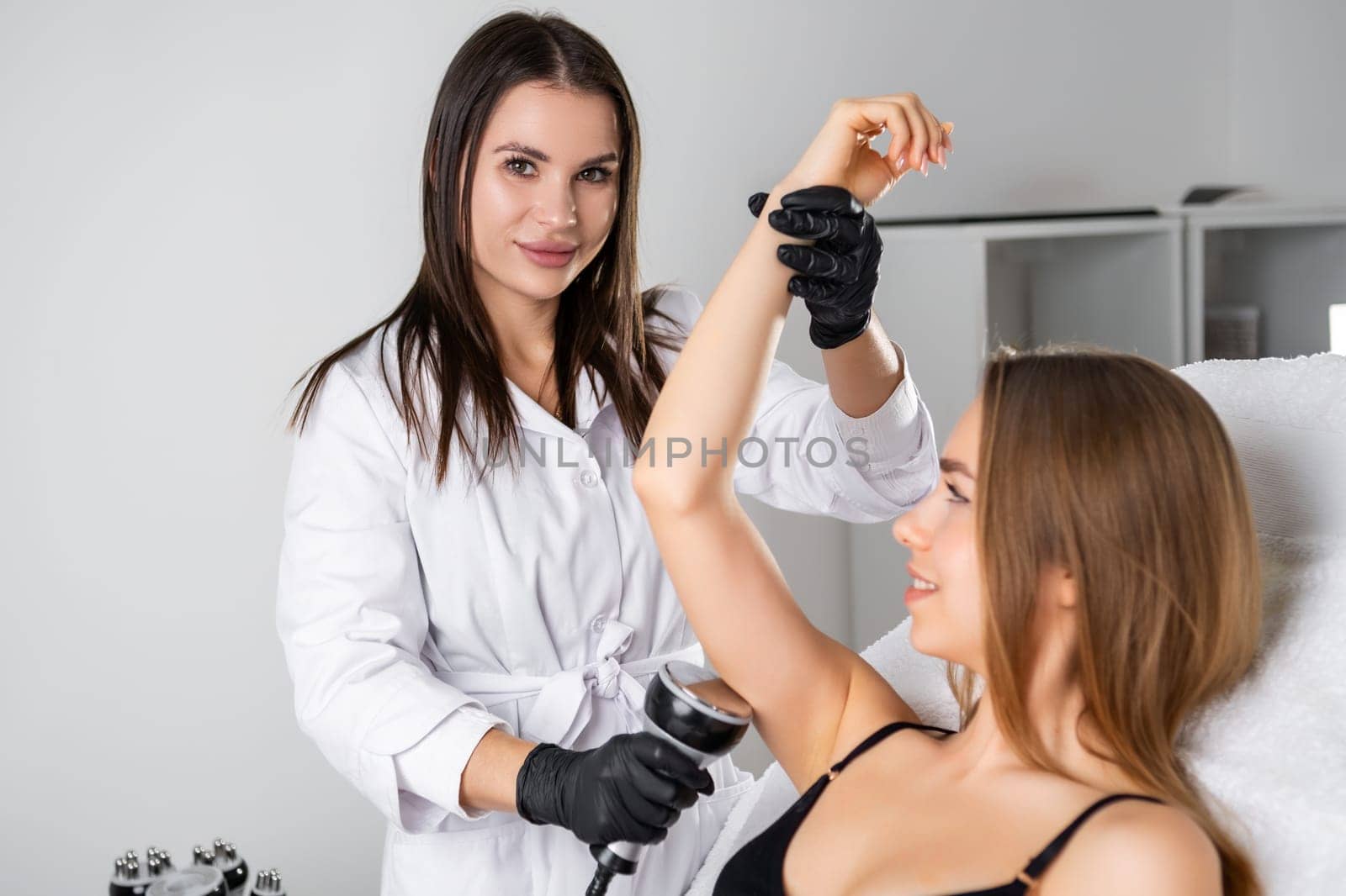Beautiful young beautician doing ultrasound cavitation on the arm of the young blonde woman.
