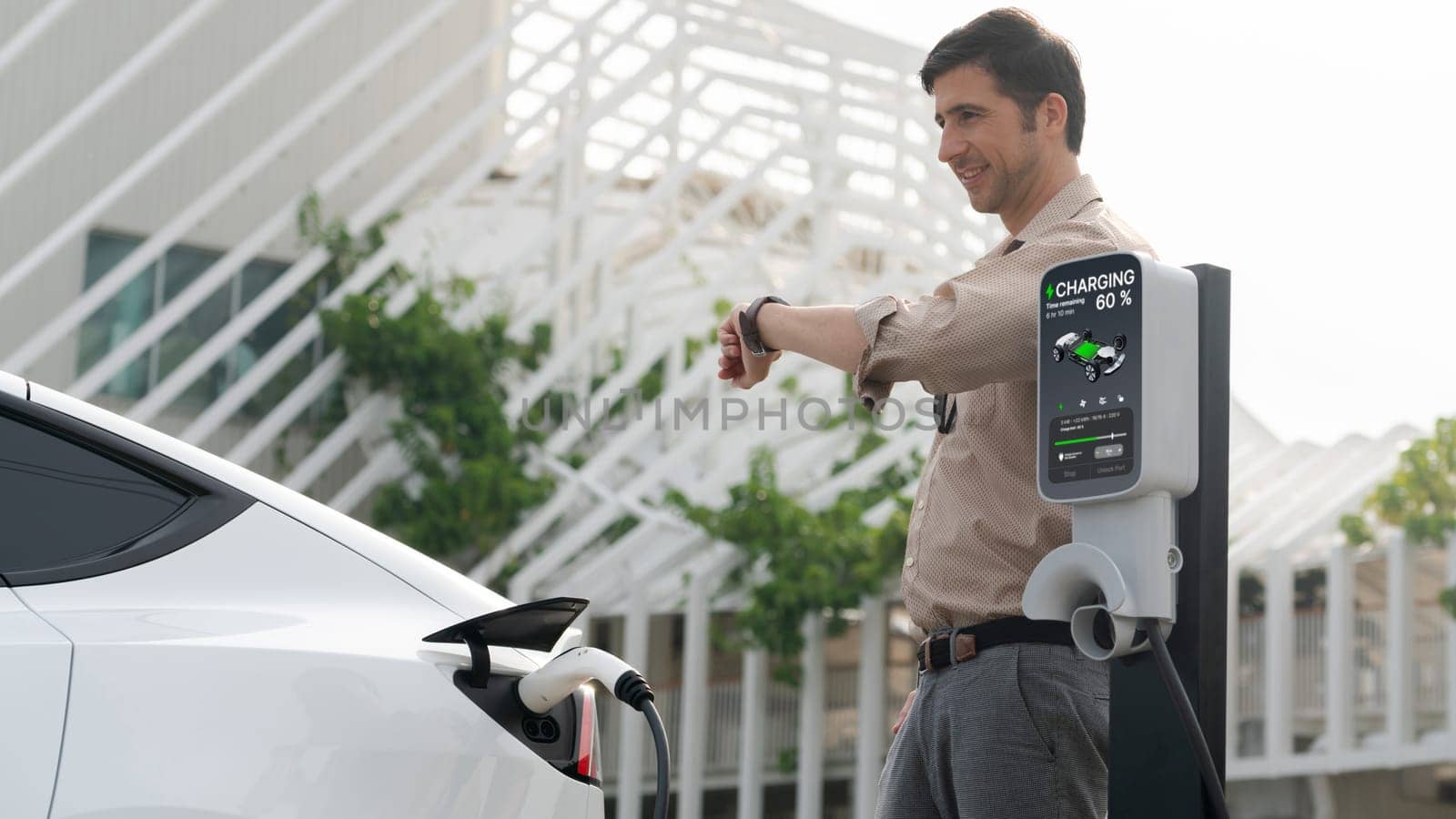 Young man checking time on smartwatch while EV charger to recharging battery from charging station in city mall parking lot. Rechargeable EV car for sustainable eco friendly urban travel. Expedient