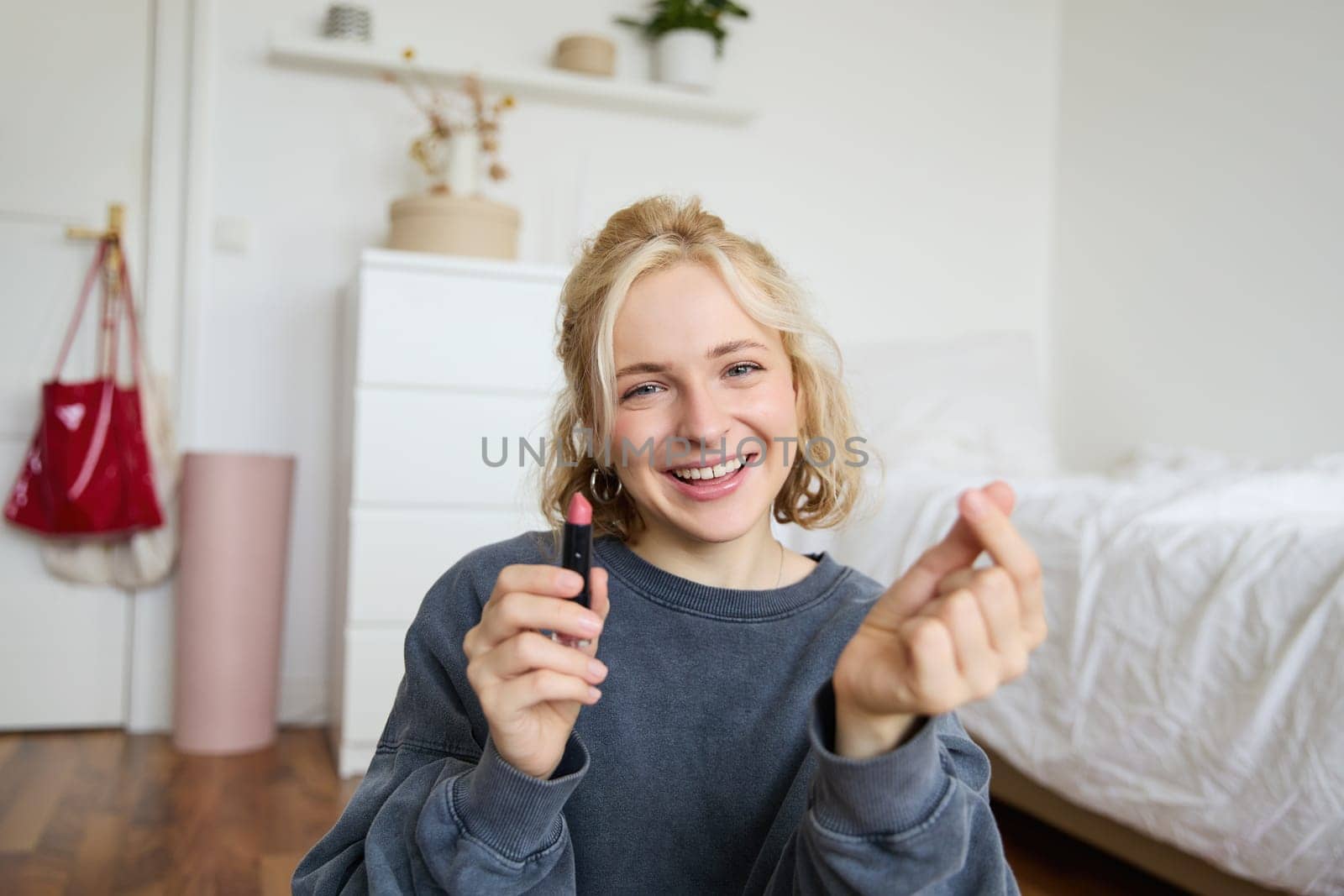 Portrait of young happy woman, content media creator recording a vlog about makeup in her room, using digital camera and stabiliser to show beauty products, holding lipstick.