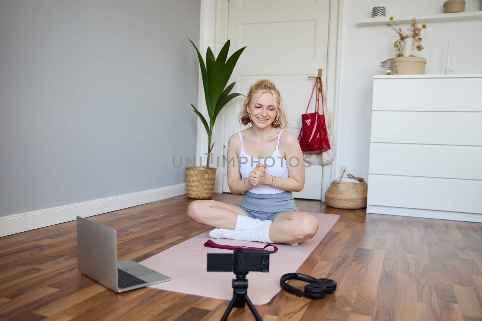 Portrait of young fitness blogger, woman showing exercises to her followers, recording video on digital camera, doing workout training session, home yoga, sitting in front of laptop on rubber mat.