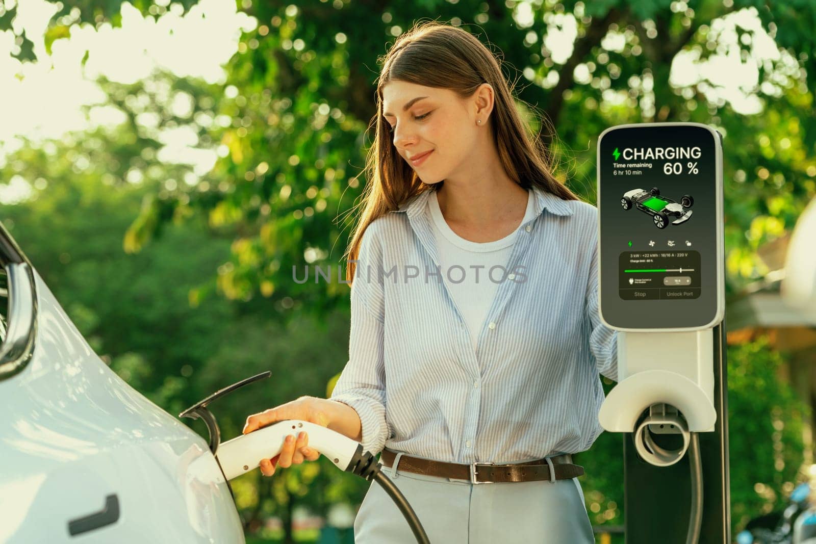 Young woman recharging battery for electric car during road trip travel EV car in natural forest or national park with sunrise lighting. Eco friendly travel during vacation and holiday. Exalt