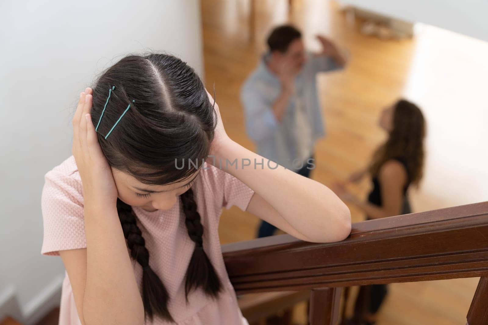 Stressed and unhappy young girl cover her ears blocking sound tension by her parent argument from the stair. Unhealthy family and domestic violence lead to traumatic childhood and anxiety. Synchronos