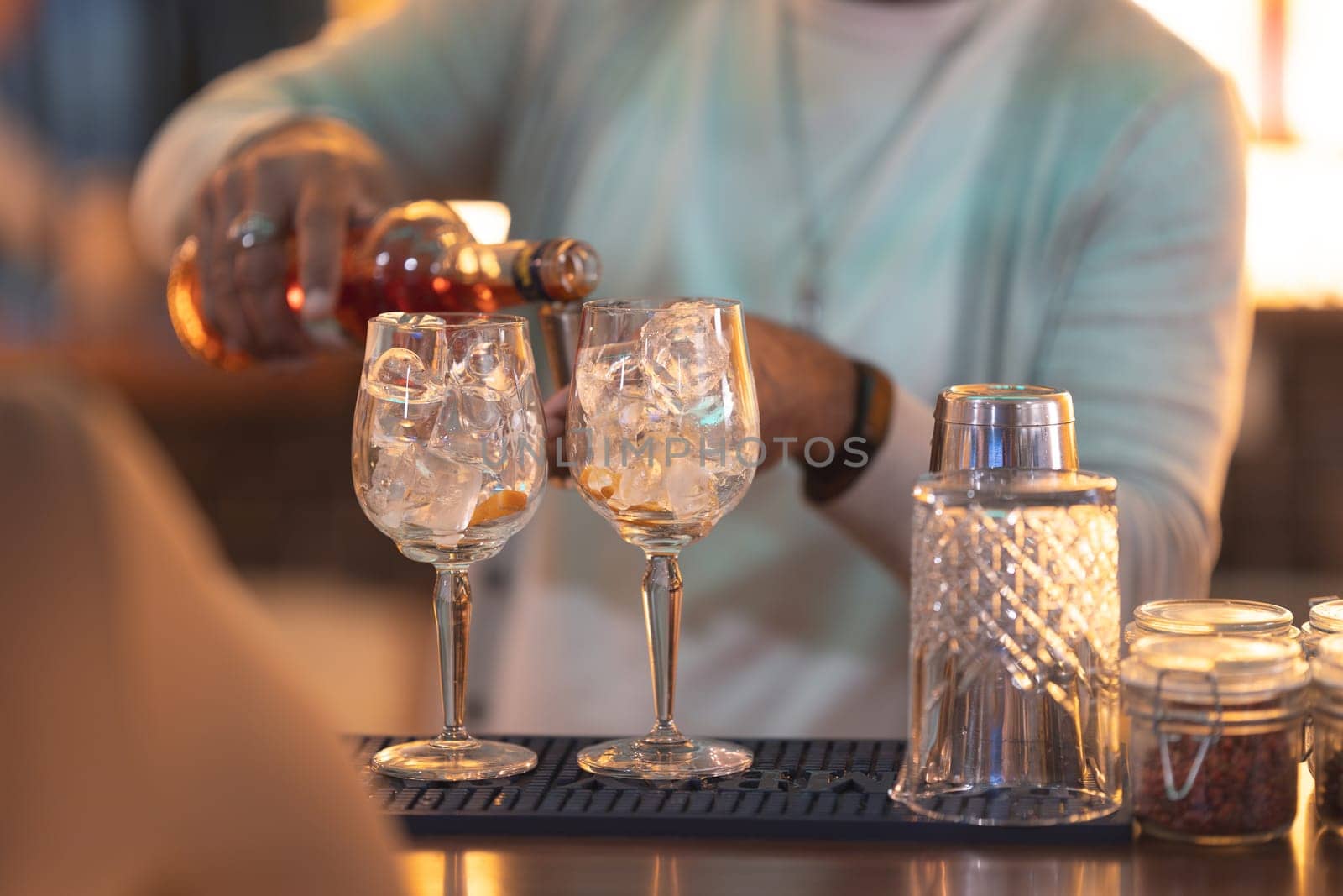 A man is pouring a drink into two glasses on a bar. The glasses are filled with ice and the drink is a mix of alcohol and soda. The man is wearing a white shirt and a blue tie