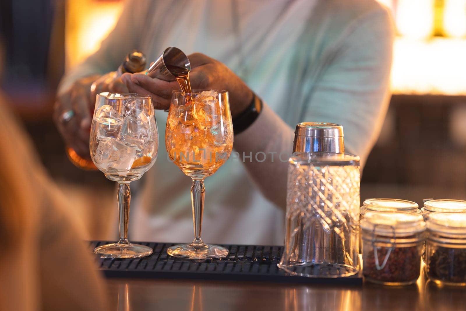 A bartender is pouring a drink into two wine glasses. The glasses are on a bar and there are other glasses and bottles around them. The bartender is wearing a white shirt and a watch