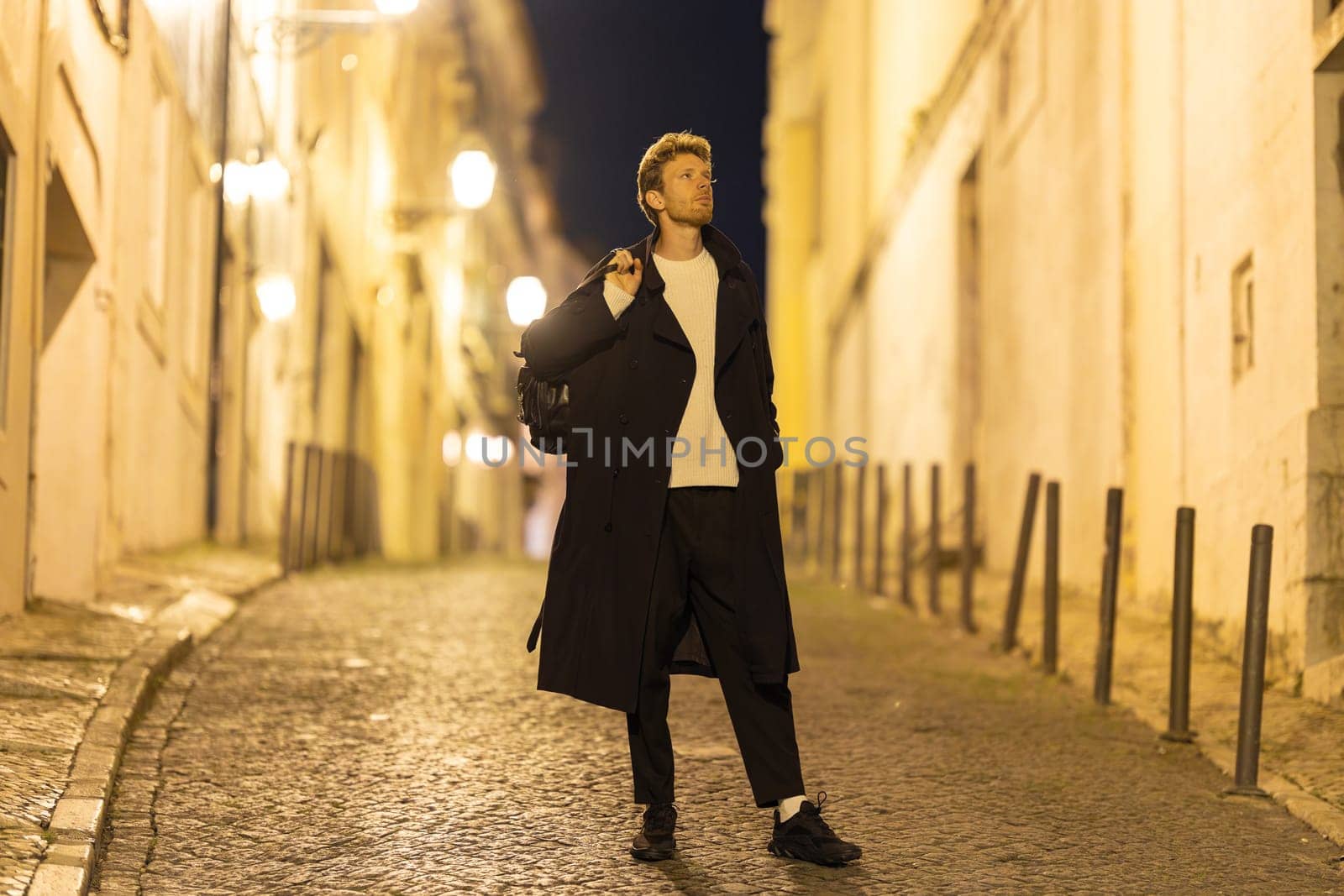 A man wearing a black coat and white shirt stands on a cobblestone street. He is holding a backpack and he is lost in thought. The street is dimly lit, giving the scene a somewhat somber