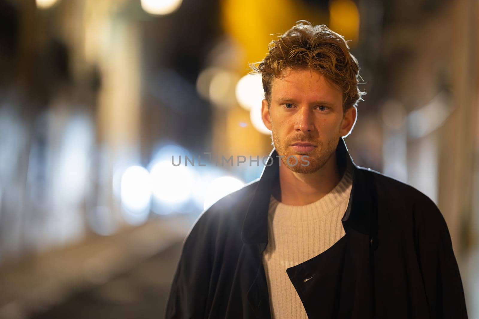 A man with red hair is walking down a street at night. He is wearing a black coat and a white shirt