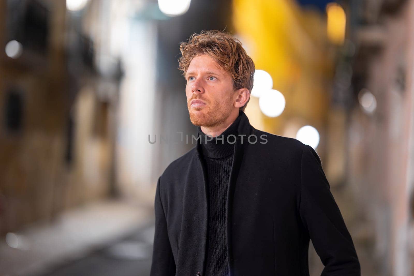 A man with red hair is standing in a city street at night. He is wearing a black coat and scarf