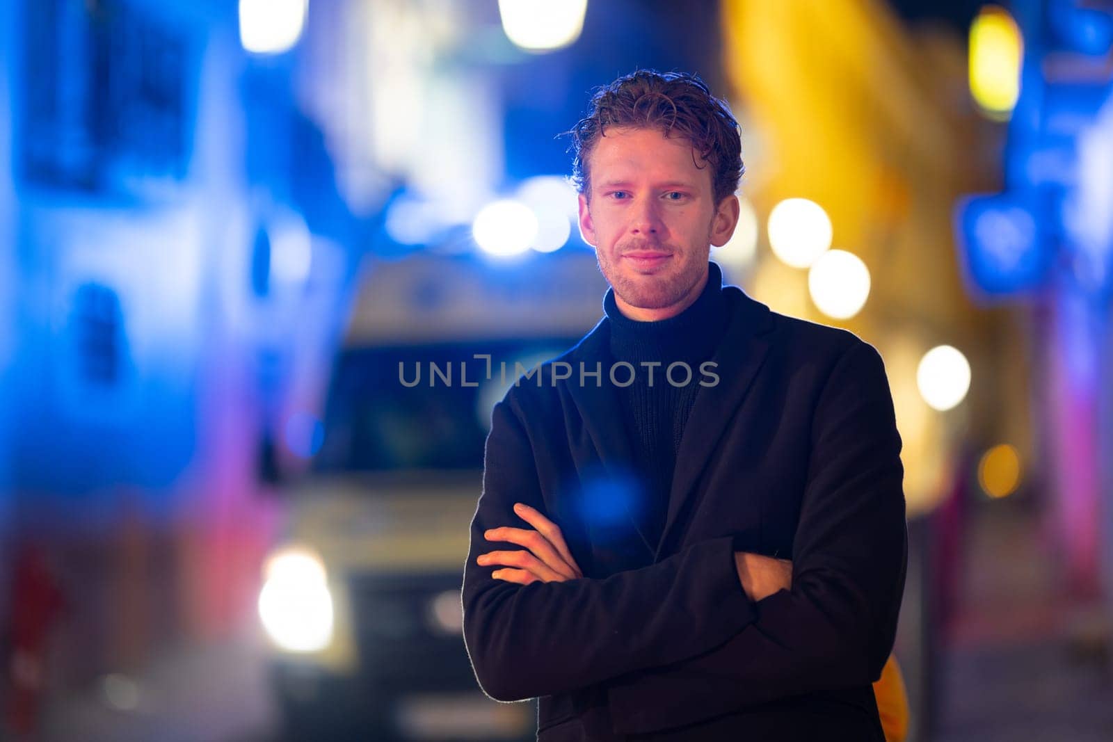 A man is standing in the street with his arms crossed. He is wearing a black jacket and a black tie. The street is lit up with bright lights, creating a moody atmosphere