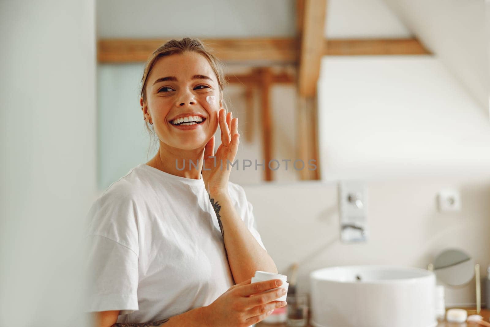Smiling woman applying hydrating moisturizer on her face sitting in bathroom. Home beauty routine by Yaroslav_astakhov
